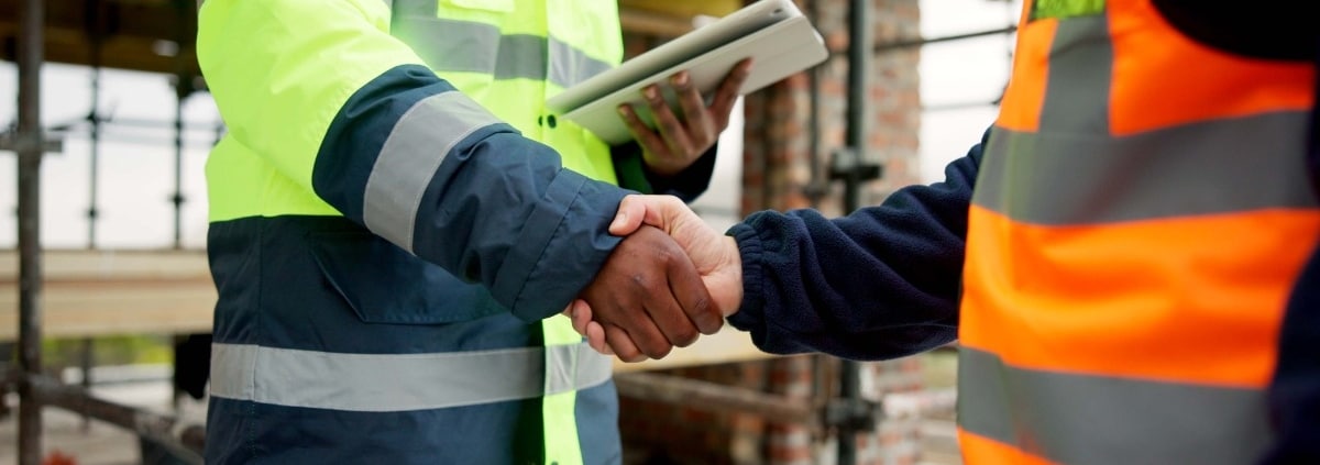 A close-up of two men shaking hands, both wearing high-visibility safety jackets, with one holding an electronic tablet.