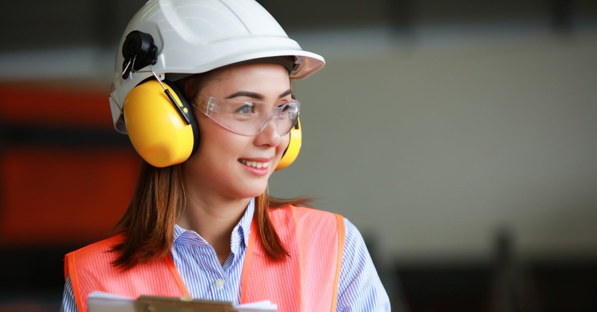 A woman wearing a hard hat, safety earmuffs, safety goggles, and a high-visibility vest holding a clipboard.