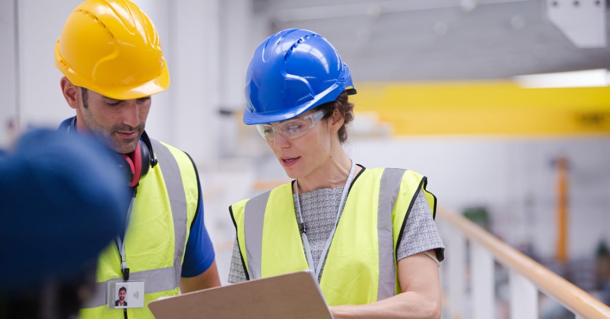 A man and a woman in high-visibility safety vests and hard hats talk while looking at a clipboard in the woman's hands