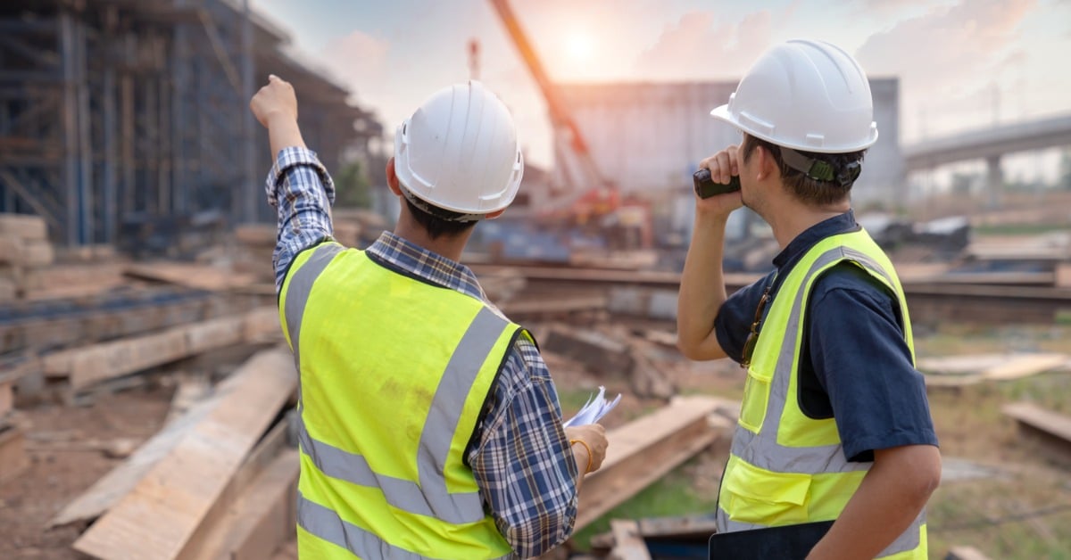 A rear view of two men in high-visibility vests and hard hats at a construction site. One is pointing toward a structure.