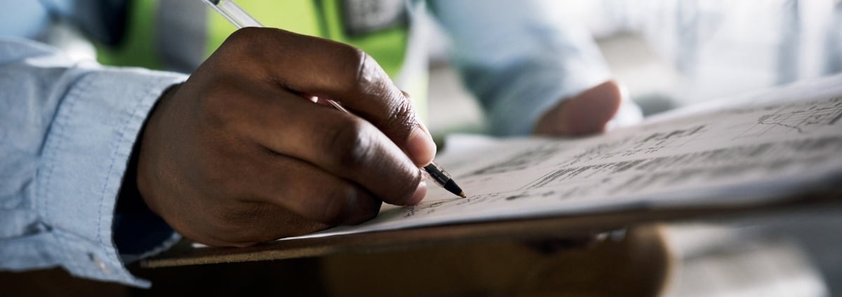 A close-up of a man writing in pen on paper attached to a clipboard while wearing a dress shirt and a high-visibility vest.