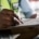 A close-up of a man writing in pen on paper attached to a clipboard while wearing a dress shirt and a high-visibility vest.