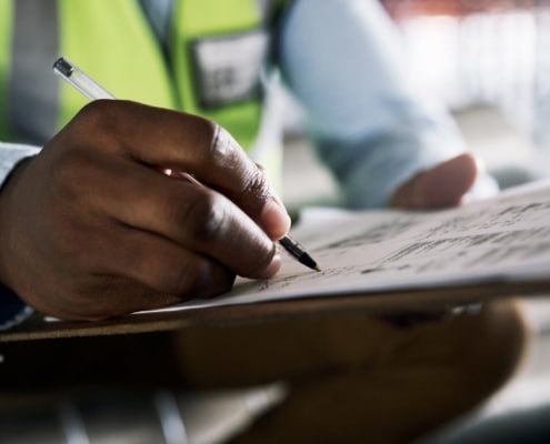 A close-up of a man writing in pen on paper attached to a clipboard while wearing a dress shirt and a high-visibility vest.
