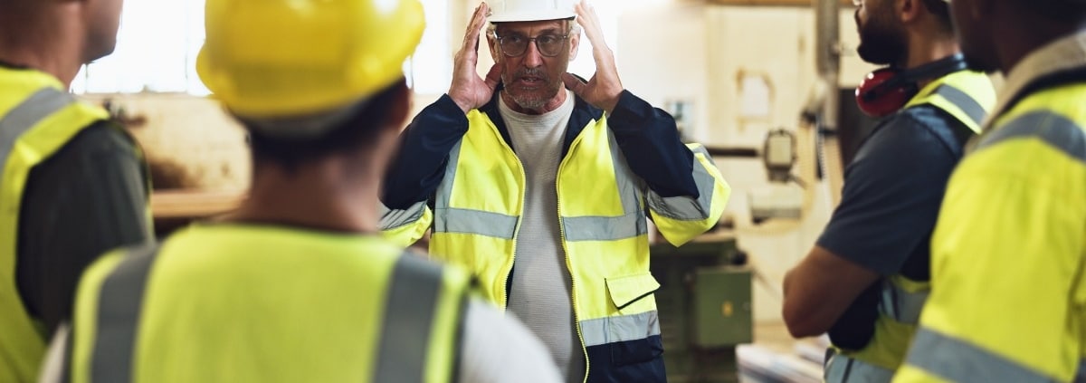 A group of men in high-visibility vests and hard hats is standing in a circle while a man touches his white helmet.