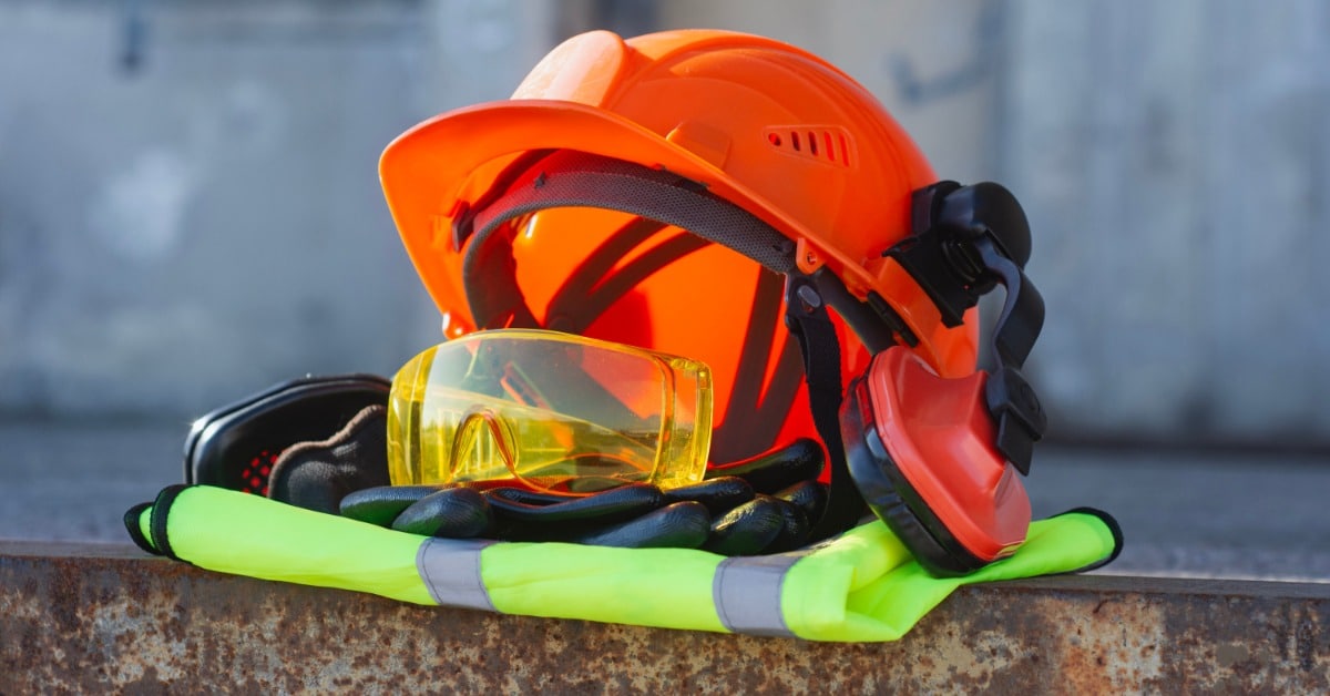 A close-up of a neat pile of construction safety gear, including a hard hat, ear protection, glasses, and a vest.