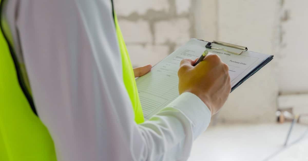 A close-up, over-the-shoulder view of a man in a dress shirt and high-visibility vest writing on a clipboard.