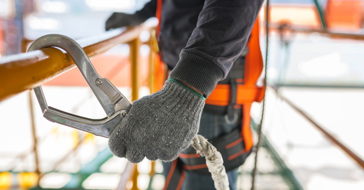 A close-up of a man's gloved hand attaching his harness to the railing of a catwalk on a construction site.