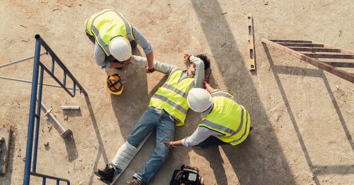 An overhead view of a construction worker on their back with their leg in a splint as other workers attend to them.