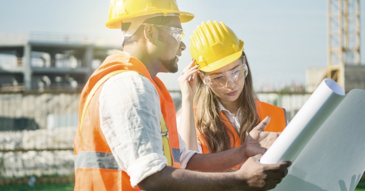 A man and a woman in high-visibility safety vests, hard hats, and safety goggles look at construction documents together.